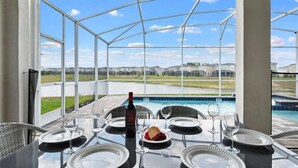 A dining area with a glass table, white plates, and wine glasses, overlooking a swimming pool and a residential area. - 9BR Lakeview Villa - Pool, Jacuzzi & Pool Table! (Kissimmee)