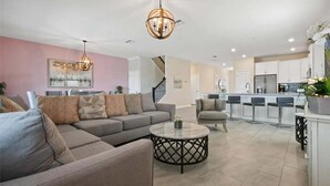 A spacious living room with a grey sofa, a round coffee table, and a chandelier. There is a kitchen area with a white countertop and bar stools. - 9BR Lakeview Villa - Pool, Jacuzzi & Pool Table! (Kissimmee)