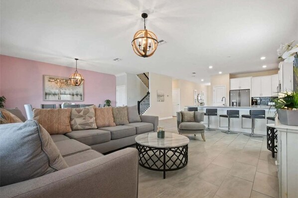 A spacious living room with a grey sofa, a round coffee table, and a chandelier. There is a kitchen area with a white countertop and bar stools.