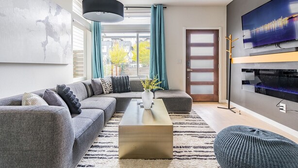 A modern living room with a sectional sofa, a coffee table, a TV mounted on the wall, and a large window with curtains. - 3BR | Rooftop | West Seattle | Sophari Home B (Seattle)