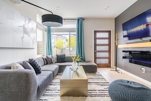 A modern living room with a sectional sofa, a coffee table, a TV mounted on the wall, and a large window with curtains. - 3BR | Rooftop | West Seattle | Sophari Home B (Seattle)