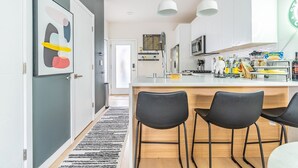 A modern kitchen with white cabinets, a central island, black chairs, and pendant lighting. - 3BR | Rooftop | West Seattle | Sophari Home B (Seattle)
