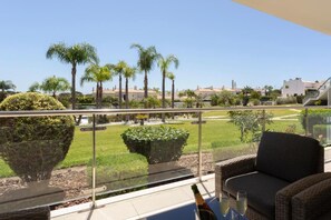 Ein Balkon mit Glasgeländer, Korbmöbeln und Blick auf einen angelegten Garten mit Palmen und einem Gebäude im Hintergrund.
