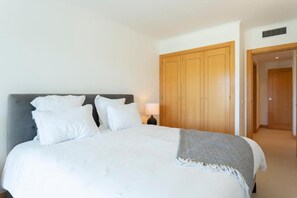 Ein ordentlich bezogenes Bett mit weißen Leinen und Kissen, eine graue Tagesdecke und ein Holeschrank im Hintergrund.