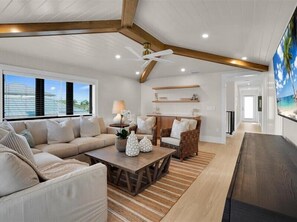 A spacious living room with a beige sofa, a wooden coffee table, and a wicker chair. There is a ceiling fan, a large window with blinds, and a TV mounted on the wall.
