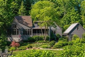 A two-story house with a porch, surrounded by lush greenery and trees. - Boathouse: Lakefront Highlands Retreat | Canoe + Gated (Highlands)