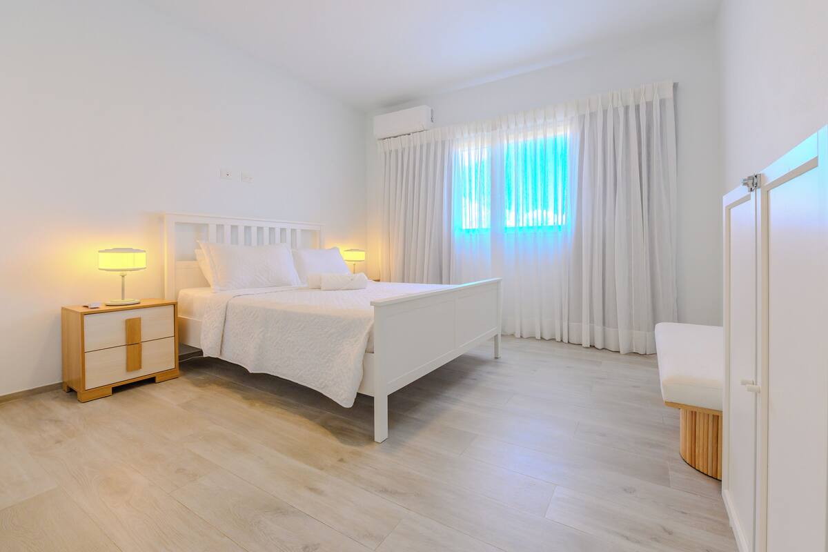 A bedroom with a bed, bedside tables, a nightstand, a window with curtains, and a door.
