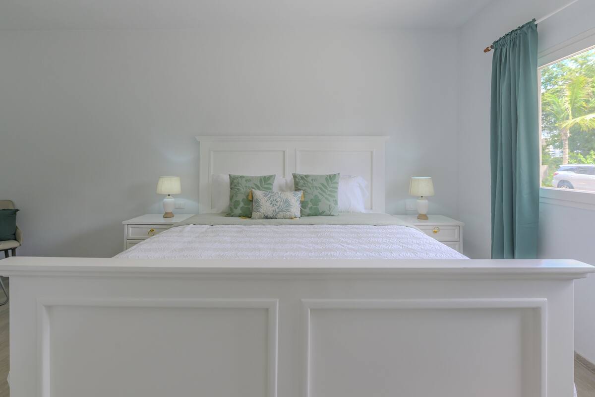 A bedroom with a white bed, two bedside tables with lamps, a window with curtains, and a view of the outside.