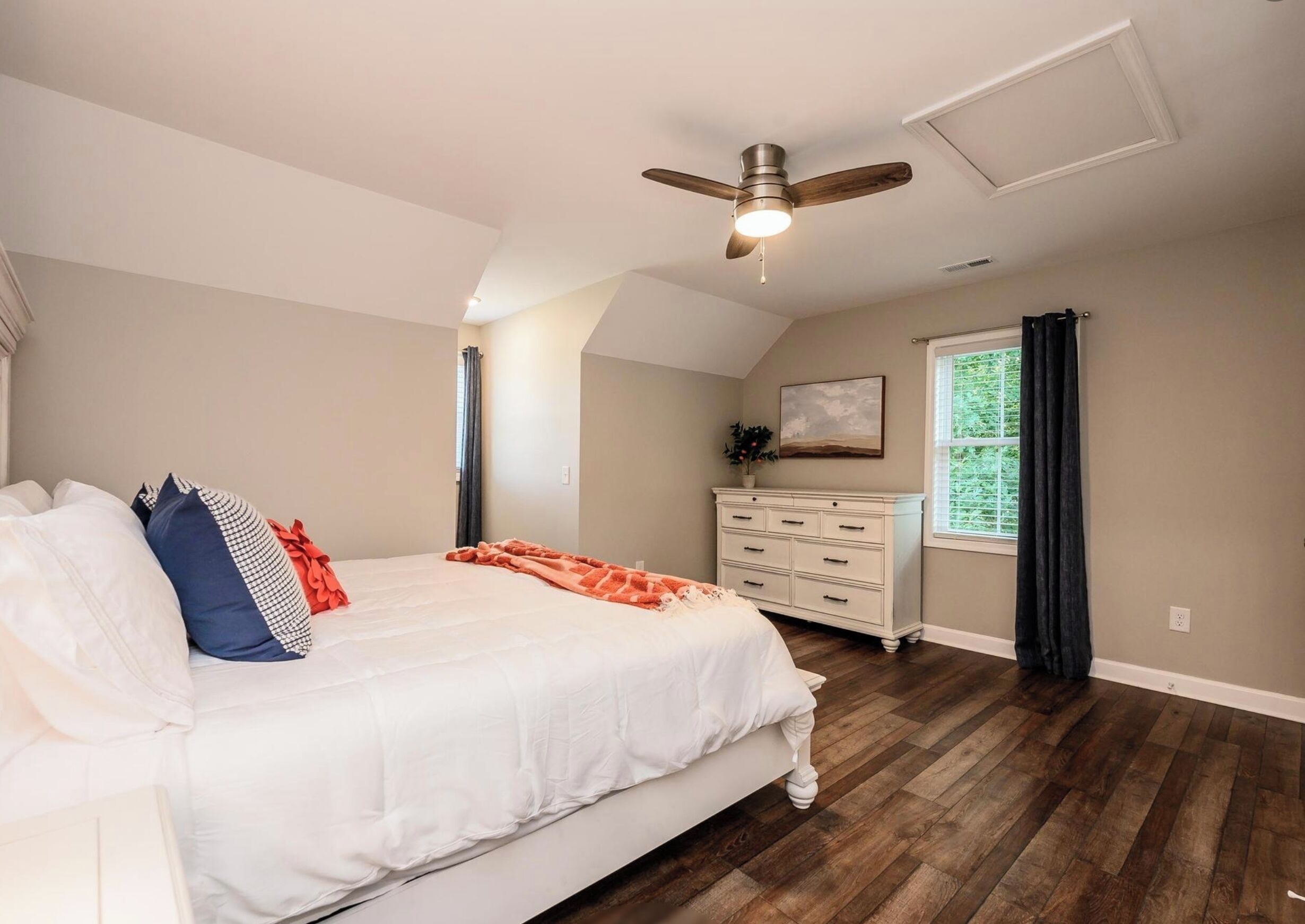 Une chambre à coucher comprenant un lit, une commode, un ventilateur de plafond et une fenêtre avec des rideaux.
