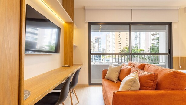 A modern living room with a large window, a flat-screen TV, a wooden desk, and an orange sofa. - Studios in Building With Gym in Brooklin - SP Emnrs (São Paulo)