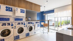 A modern laundry facility with three SEGA washing machines, a wooden panel wall, and a seating area with a view of the city. - Studios in Building With Gym in Brooklin - SP Emnrs (São Paulo)