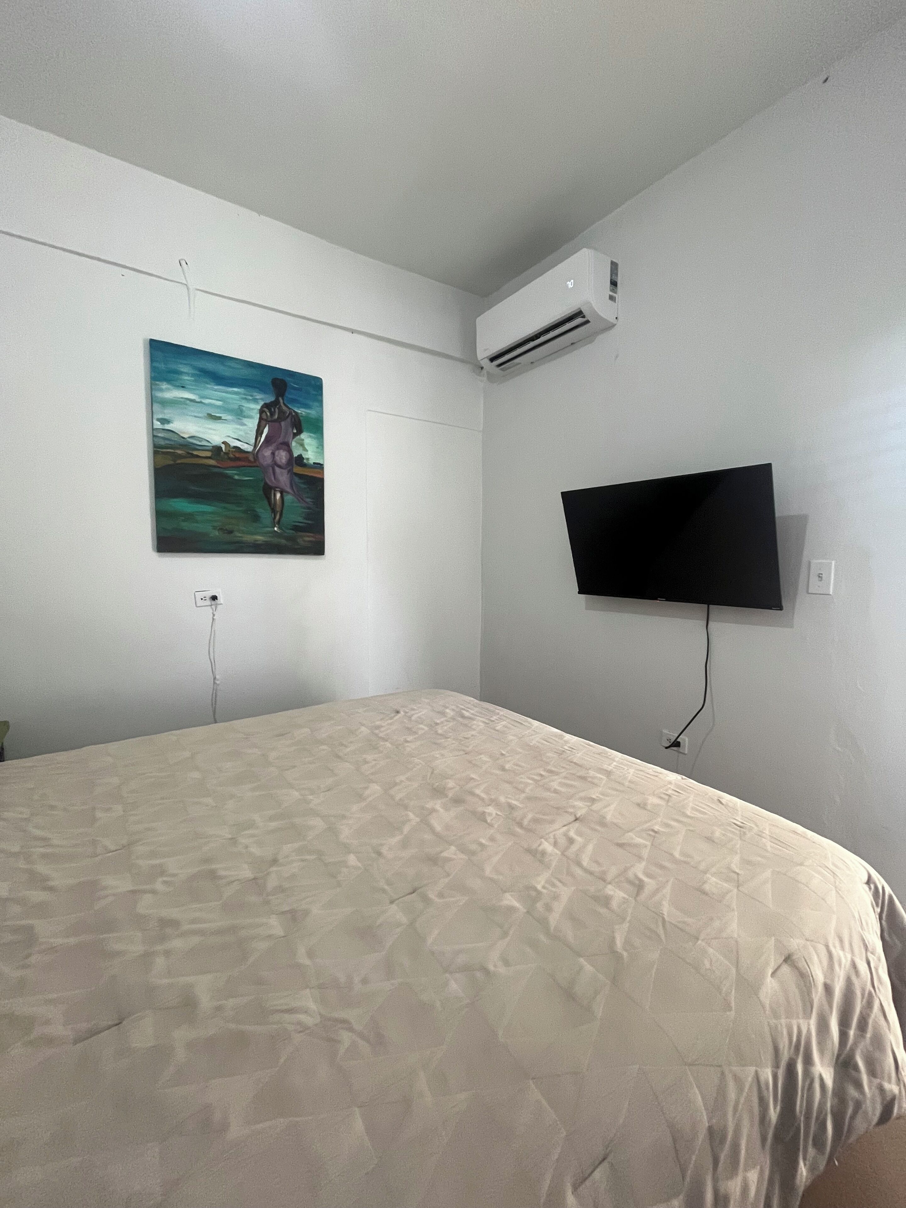 A bedroom with a bed, a painting, an air conditioner, and a TV mounted on the wall.