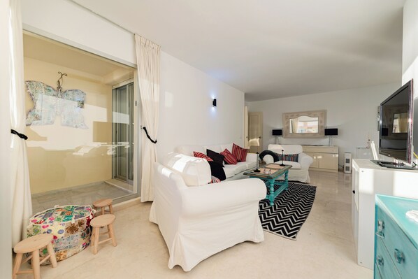 Een moderne woonkamer met een witte bank, een klein bijzettafeltje en een televisie. Er is een schuifdeur naar een balkon met een gordijn met een patroon.
