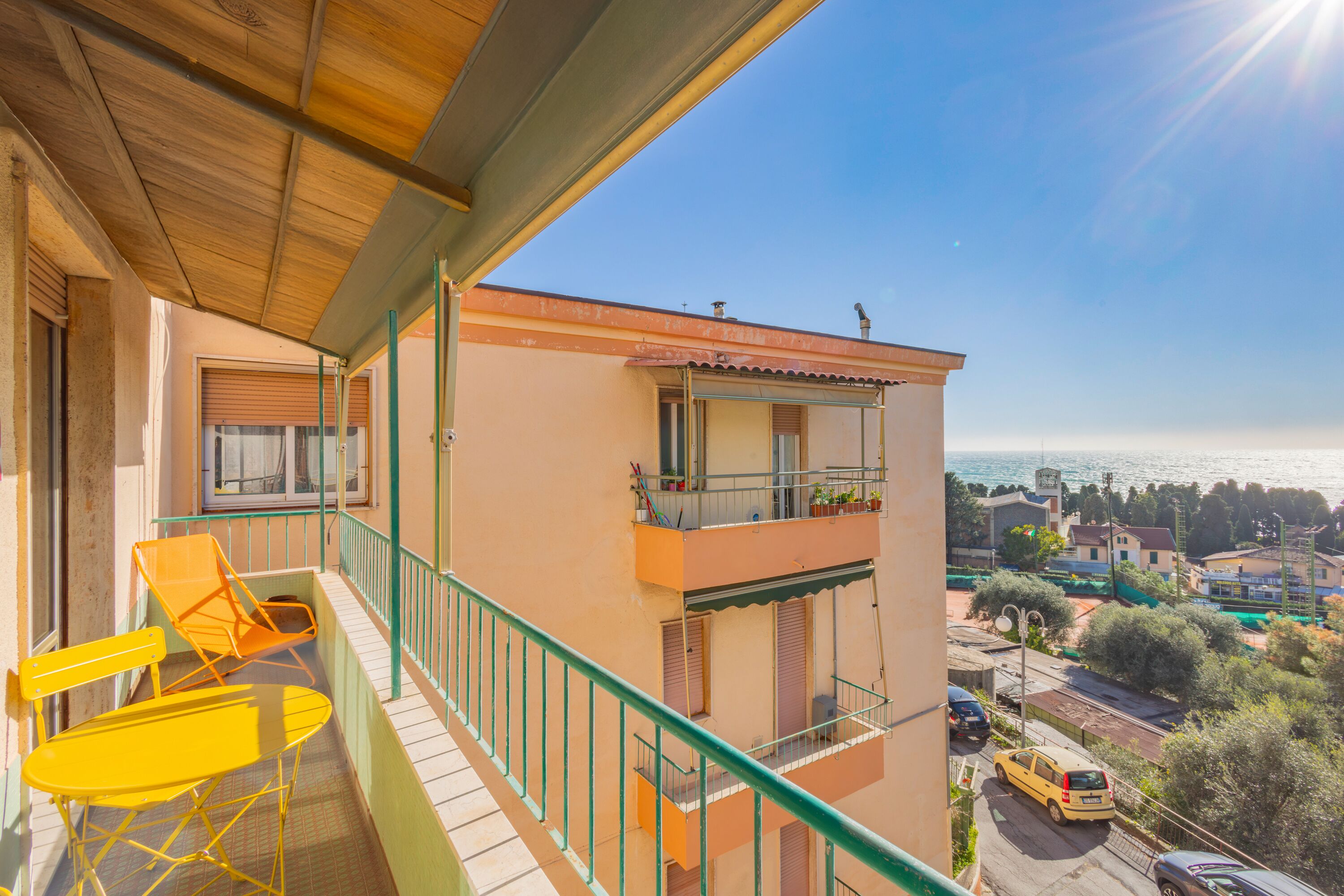 A balcony with yellow chairs and a table, overlooking a street and buildings with a view of the sea.