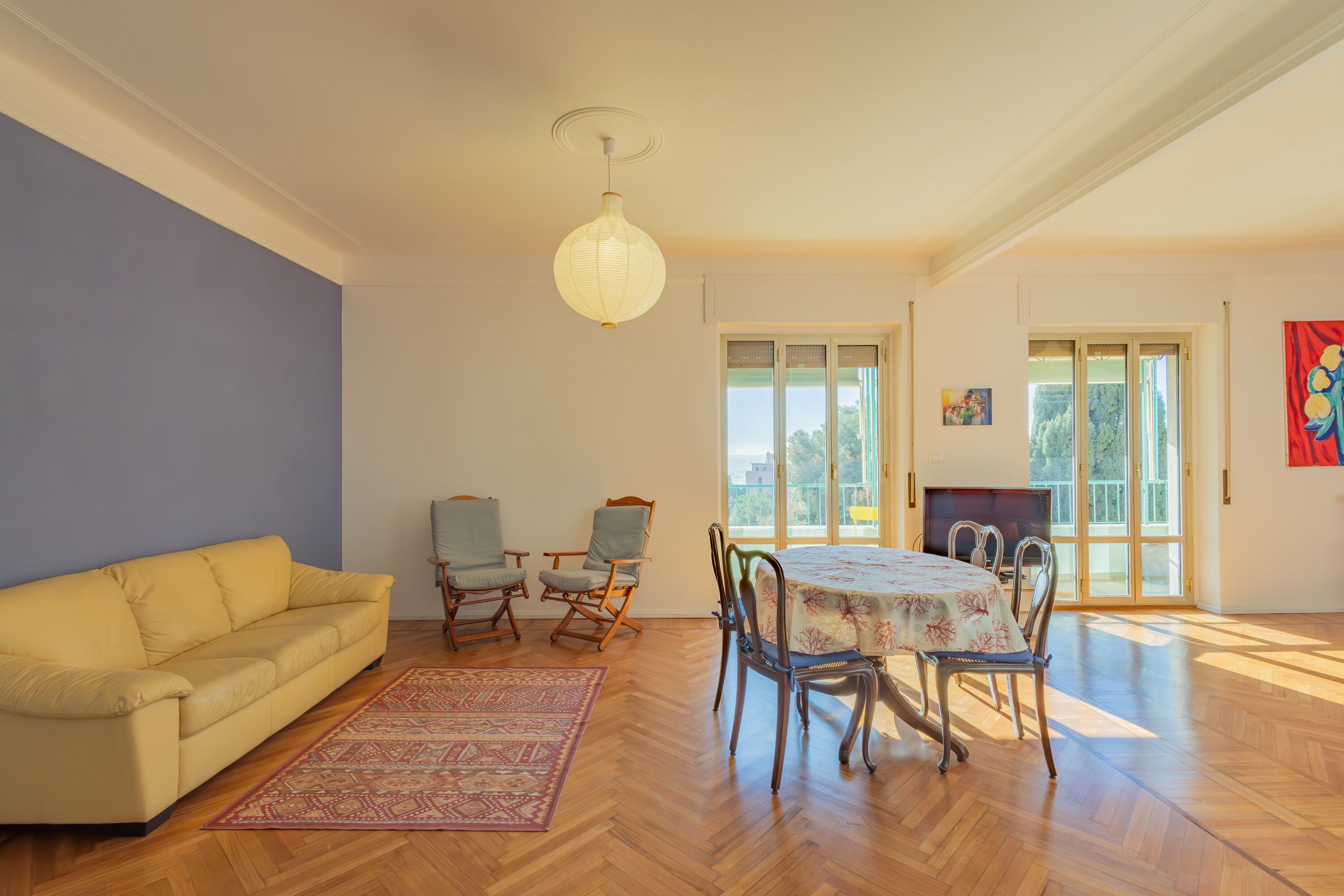 A living room with a yellow sofa, a dining table with chairs, a patterned rug, and a hanging light fixture.