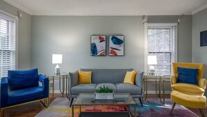 En stue med en grå sofa, blå og gule lænestole, et glasbord og abstrakt kunst på væggen.