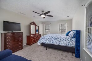Une chambre à coucher comprenant un lit, une commode, un téléviseur, un ventilateur de plafond et des fenêtres.