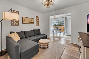 A modern living room with a grey sectional sofa, a round ottoman, a floor lamp, and a dining area with a table and chairs. - Urban Suite | An Inviting 1-Bedroom City Escape (Temple)