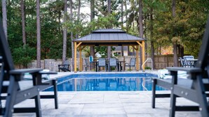 A swimming pool with a gazebo, surrounded by chairs and tables, set in a wooded area. - Luxury Golf Retreat - Simulator & Short Game Setup (Southern Pines)