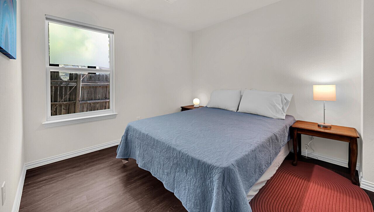 A bedroom with a bed, a nightstand, a lamp, a window, and a view of a wooden fence.