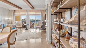 Un vaste salon doté d’un haut plafond, de grandes fenêtres et offrant une vue sur l’extérieur. On y trouve plusieurs coins salon avec des meubles confortables, ainsi qu’une étagère décorative où sont exposés divers objets.