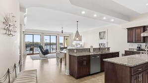 Een ruime keuken met donkerhouten kastjes, granieten aanrechtbladen en een groot eiland. Er is een zithoek met een stoel en een bank, en uitzicht op de omgeving door grote schuifdeuren.