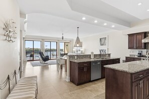 A spacious kitchen with dark wood cabinets, granite countertops, and a large island. There is a dining area with a chair and a bench, and a view of the outdoors through large sliding doors. - Gulf-Front Luxury | Family Friendly | 3-Bedroom Escape (Santa Rosa Beach)