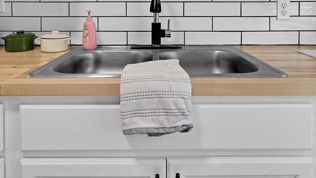 Una cocina con armarios blancos, un lavamanos de acero inoxidable, una olla verde, una olla blanca, una botella rosa y una toalla colgada en la puerta.