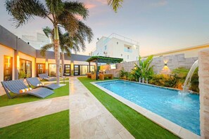 A modern outdoor pool area with lounge chairs, a small seating area, and a waterfall feature. - New Sea La Vie Suite # 3 |2BR|1BR W. Pool&Jacuzzi (Noord)
