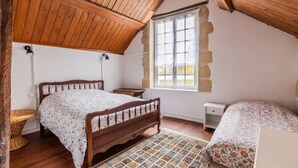 Une chambre avec un plafond en bois, un lit recouvert d’une couvre-lit à motifs floraux, une fenêtre encadrée de murs en pierre et une petite table de chevet.