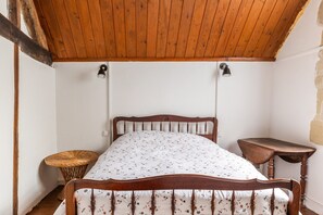 A wooden ceiling, a bed with a floral bedspread, a wicker chair, and a small wooden table. - Holiday Home “La Maison Blanche” with Private Terrace and Wi-Fi (Nazelles-Négron)