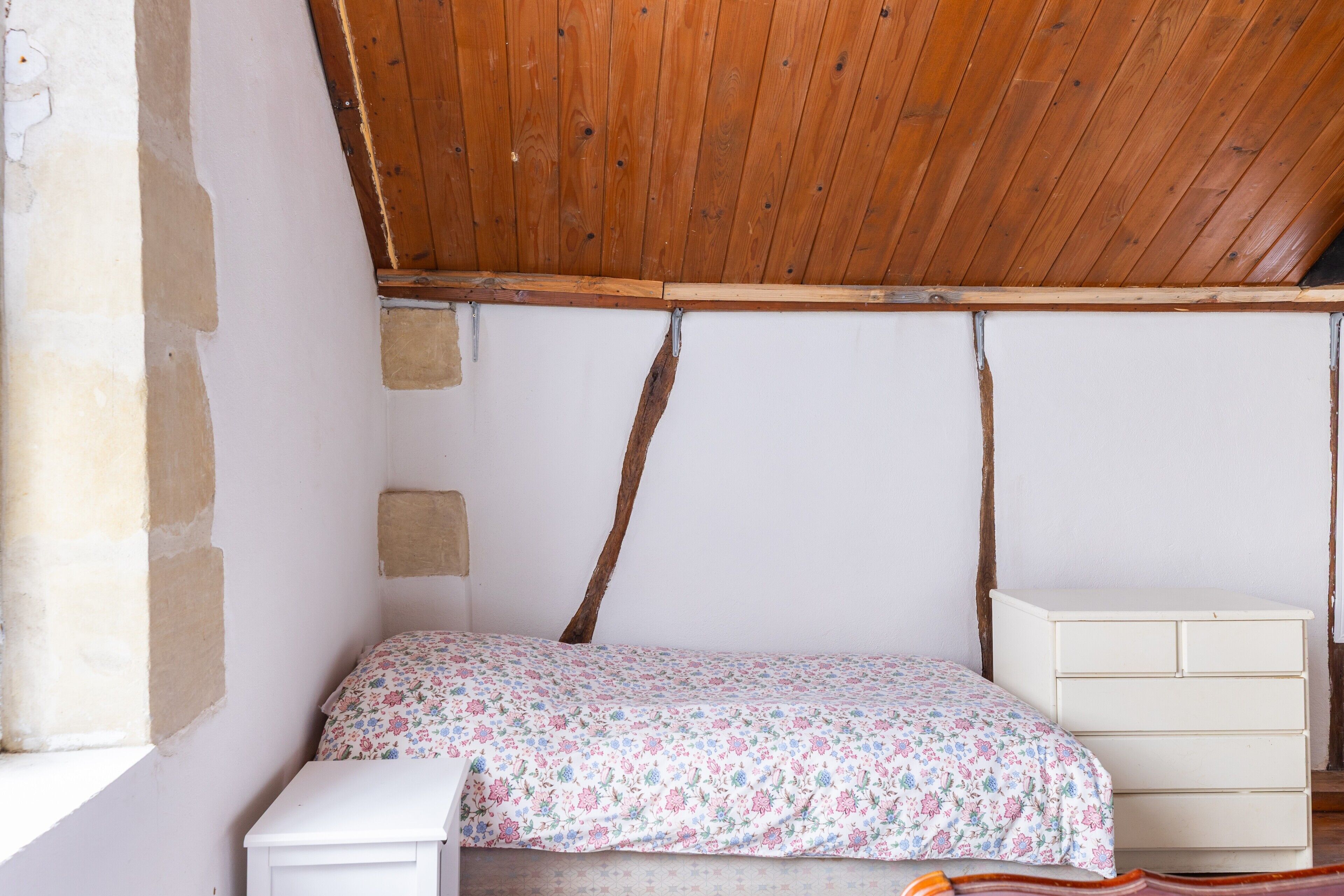 Un dormitorio con techo de madera, una cama con colcha floral, un aparador blanco y una ventana.