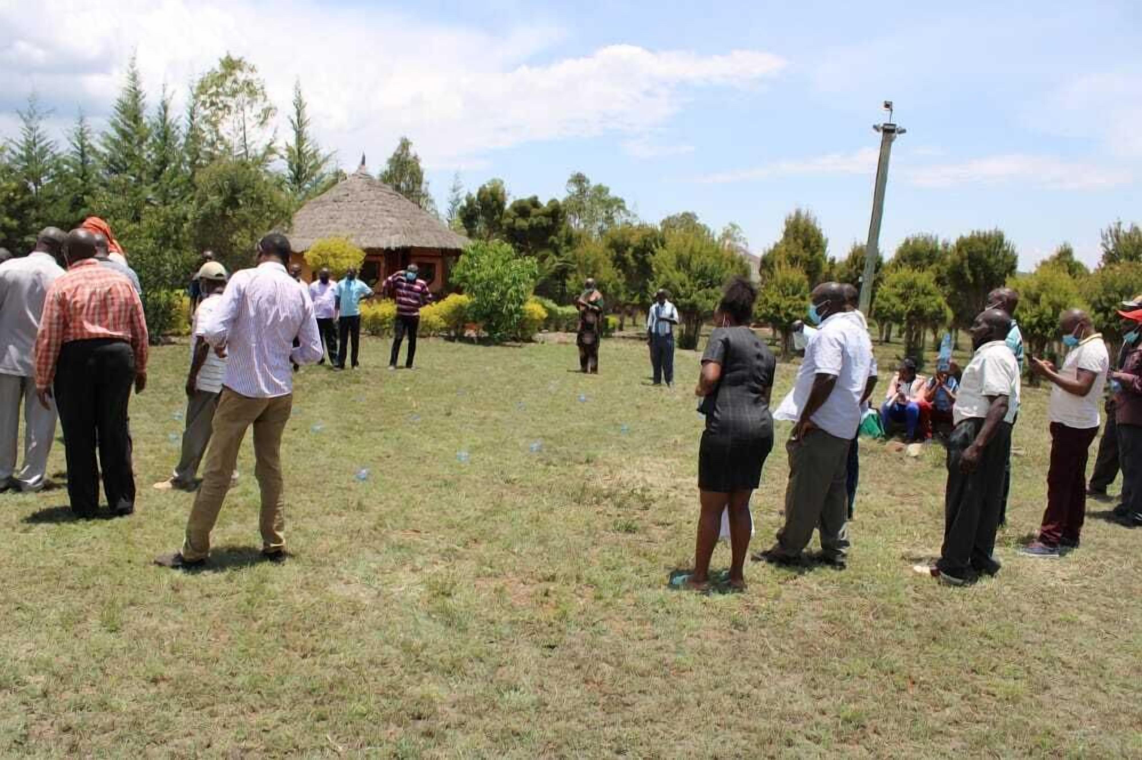 Un grupo de personas reunidas al aire libre en un área con césped, árboles y una construcción con techo de paja al fondo.