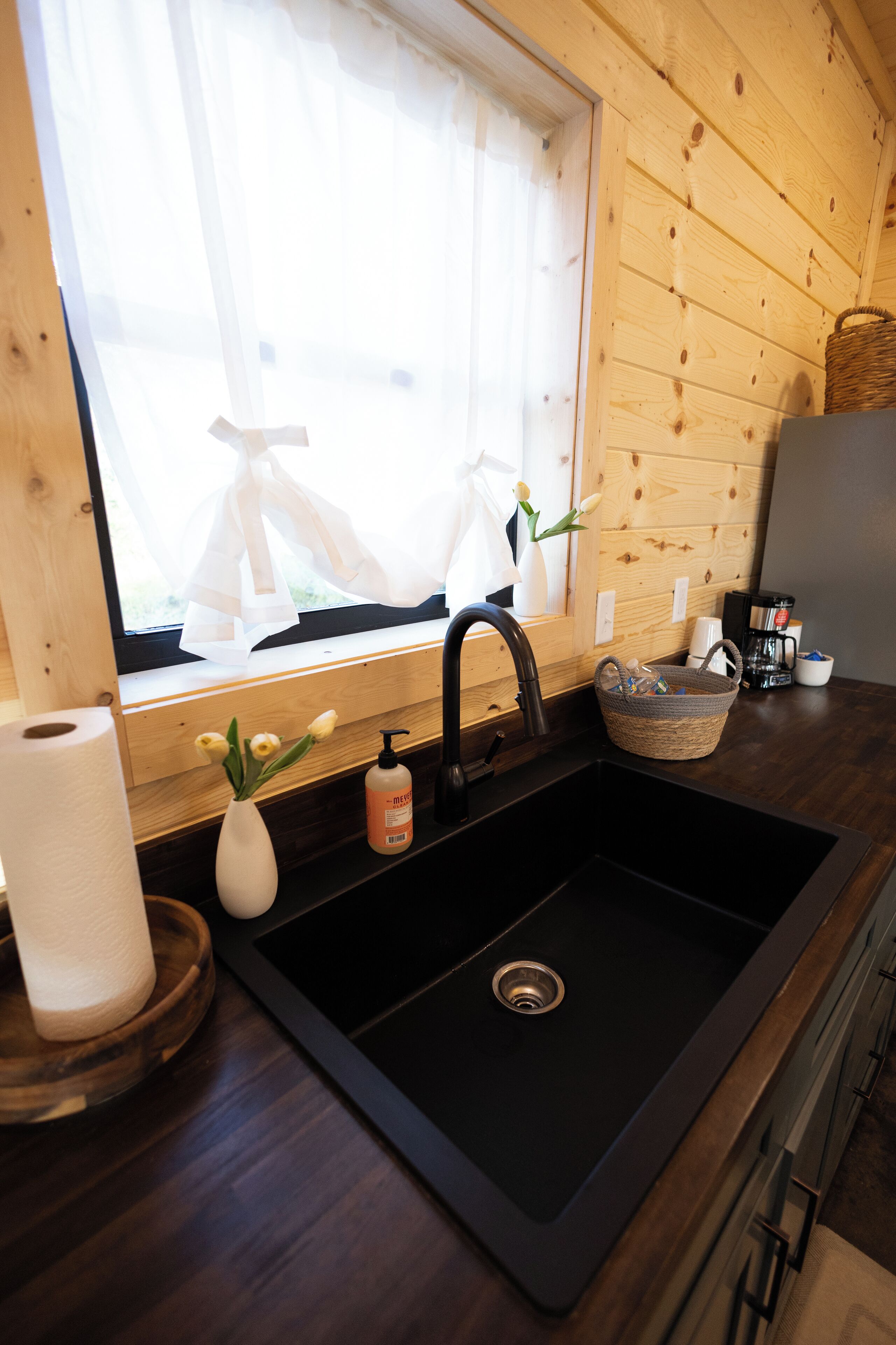 A kitchen with a black sink, wooden walls, a window with sheer curtains, a vase with flowers, and a basket on the counter.