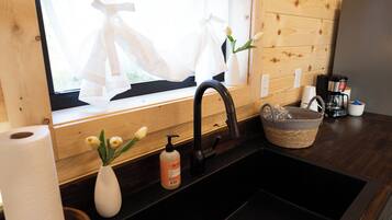 A kitchen with a black sink, wooden walls, a window with sheer curtains, a vase with flowers, and a basket on the counter.