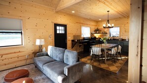Ein gemütliches Blockhäuschen-Interieur mit einer Couch, Poufs, einem Esstisch mit Stühlen, einer Küchenzeile und einem großen Fenster.