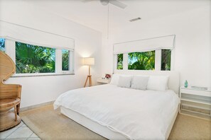 A bedroom with a large bed, a chair, a lamp, and a window with a view of palm trees. - Ground Floor Yacht Club Condo E1 - Sunset Views, BBQ, Walk to Beach and Dining (Providenciales)