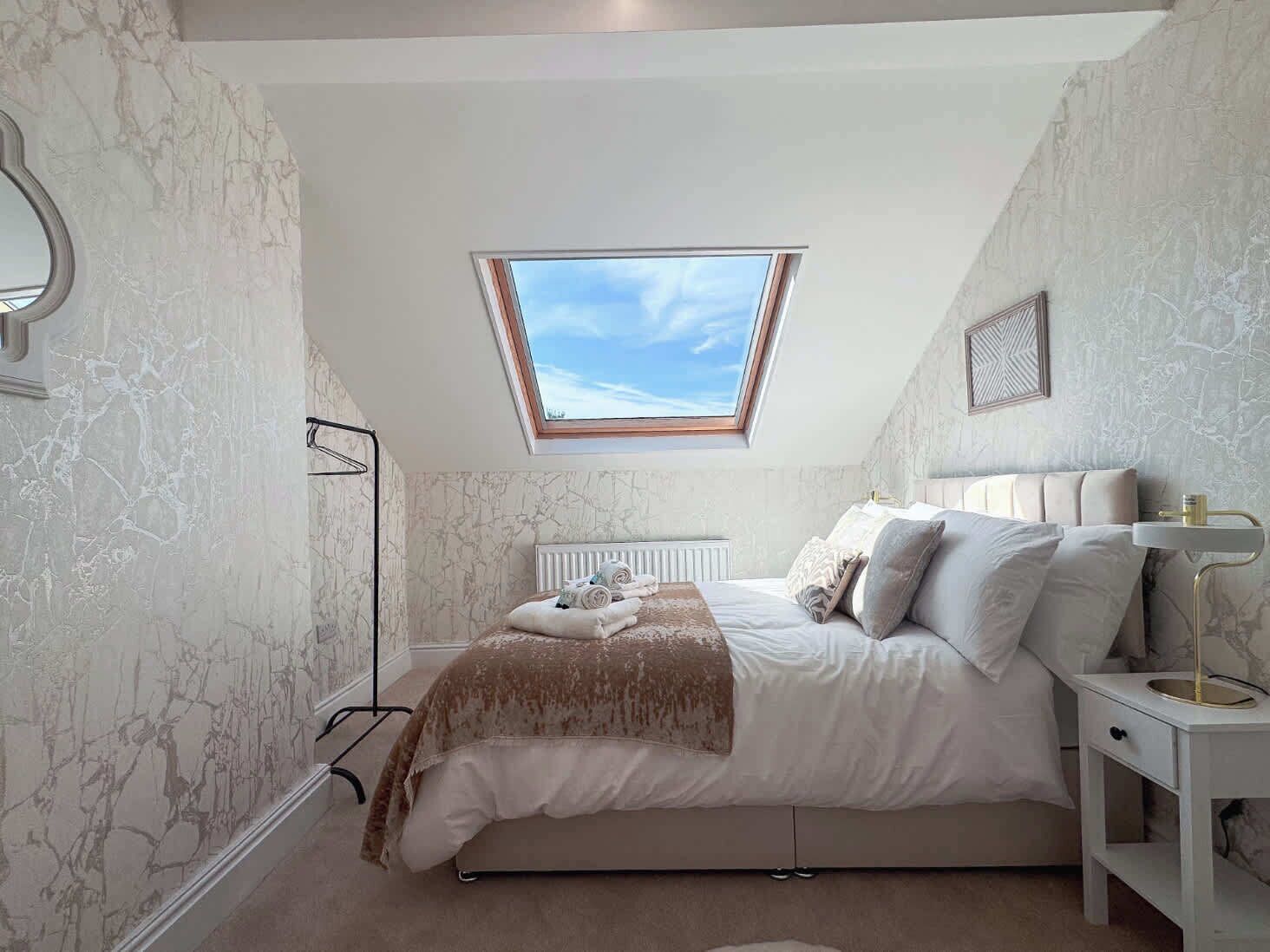 A bedroom with a skylight, a bed with white bedding, a bedside table with a lamp, and marble-patterned walls.