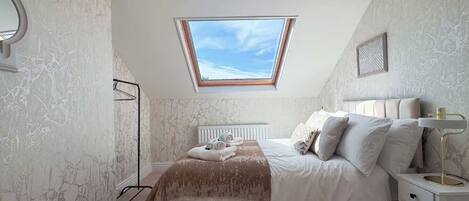 A bedroom with a skylight, a bed with white bedding, a bedside table with a lamp, and marble-patterned walls.