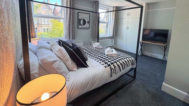A modern bedroom with a large bed, a TV, and a view of buildings through large windows. - 6 Bedroom Victorian Townhouse in Central Harrogate (Harrogate)