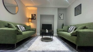 A modern living room with two green sofas, a white rug, a black coffee table, a flat-screen TV, and a fireplace. - 6 Bedroom Victorian Townhouse in Central Harrogate (Harrogate)