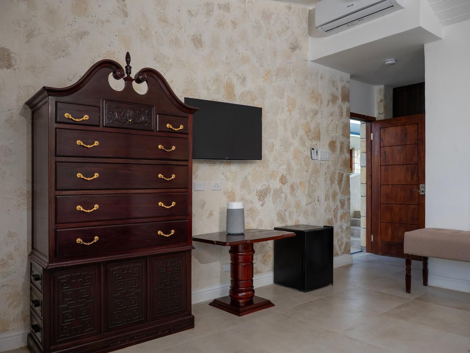 A room with a large wooden dresser with gold handles, a flat-screen TV mounted on the wall, a small table with a candle, and a wooden door.