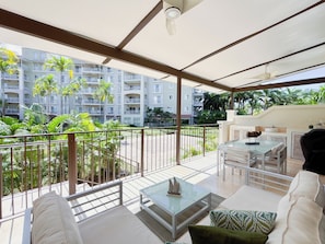 A balcony with a glass table, white sofa, and a view of a multi-story building with balconies and palm trees.