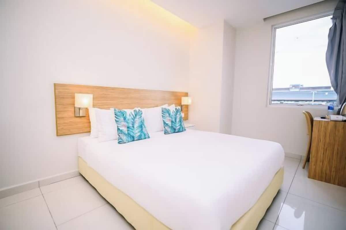 Ein Hotelzimmer mit einem großen Bett, zwei Wandlampen, einem hölzernen Kopfteil und einem Fenster mit Blick auf die Stadt.