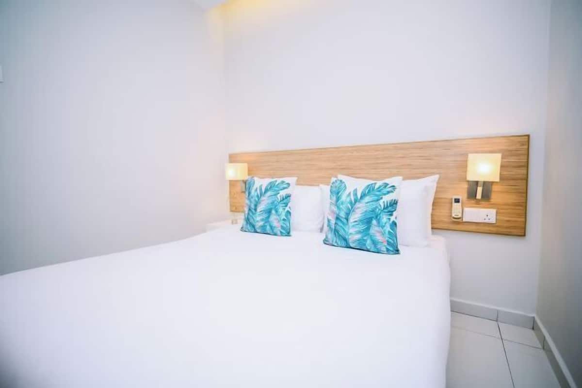 Ein Hotelzimmer mit einem Holzkopfteil, zwei Wandlampen und einem Bett mit weißer Bettwäsche und Kissen mit blauem Blattmuster.