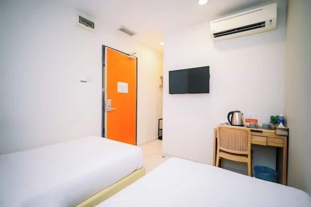 Ein Hotelzimmer mit zwei Einzelbetten, einem Schreibtisch mit Stuhl, einer orangen Tür, einem flachbildfernseher an der Wand und einer Klimaanlage.
