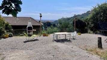 Eine gepflasterte Terrasse mit Tisch und Stühlen, einem Grill und Blick auf eine Hügellandschaft mit Häusern.