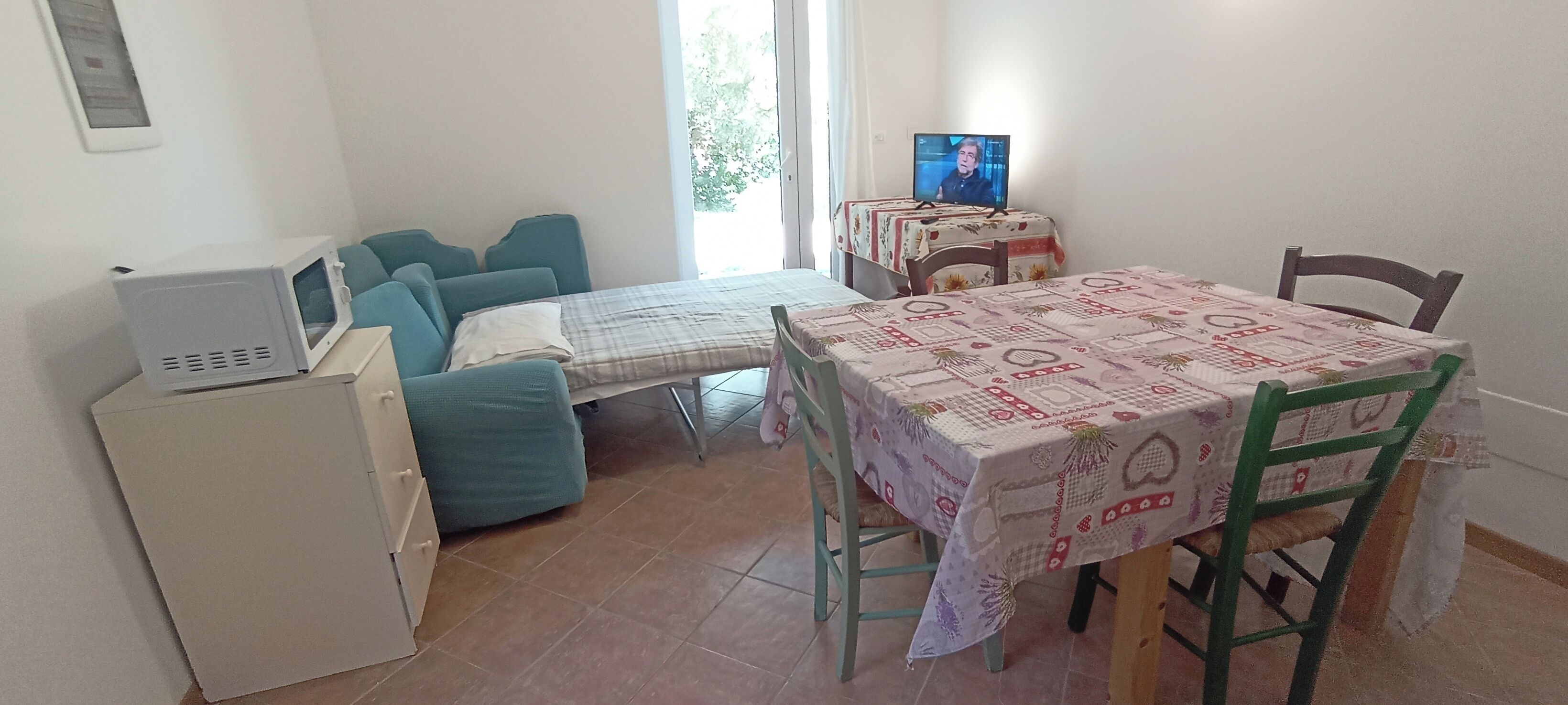A small room with a bed, a table with a tablecloth, a television, and a microwave.