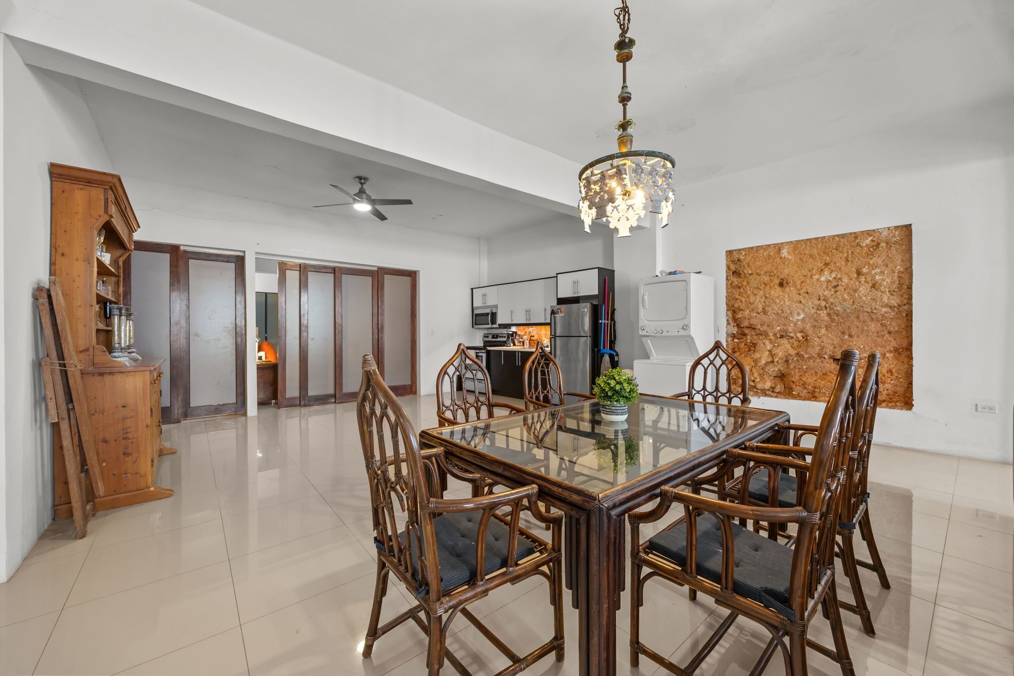 A spacious dining area with a glass-top table surrounded by wooden chairs, a chandelier, and a modern kitchen in the background.