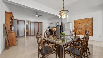 A spacious dining area with a glass-top table surrounded by wooden chairs, a chandelier, and a modern kitchen in the background.